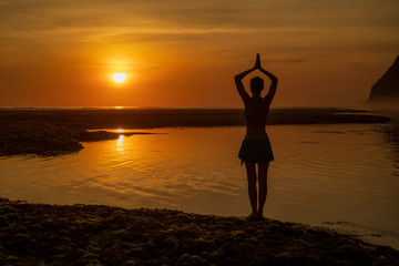 Yoga pose. Woman standing on the beach, practicing yoga. Young woman raising arms with namaste mudra during sunset golden hour. View from back. Melasti beach, Bali.