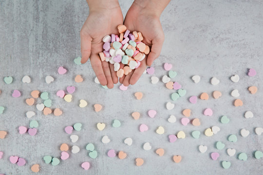 Valentine's Day hands holding conversation heart candies and scattered conversation hearts scattered on a gray cement neutral background