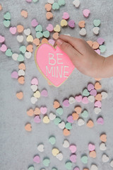 Fototapeta premium Valentine's Day hand holding a be Mine Heart Shaped pink sugar cookie and scattered conversation hearts scattered on a gray cement neutral background