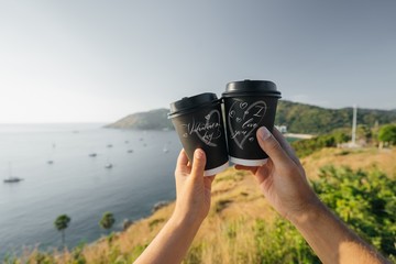 Couple of lovers hold in their hands paper cups with coffee, on a background of mountains and the sea. On the cups there is the inscription "St. Valentine's Day", "I love you."