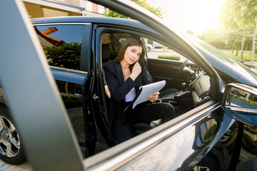 Young businesswoman talking on the phone in the passenger seat of the car and holding in hand a clipboard with paper for writing notes