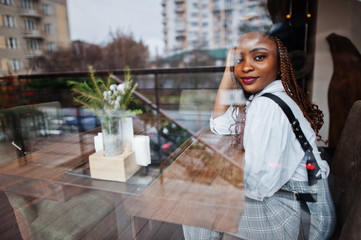 African american woman in overalls and beret sitting on cafe against glass window.