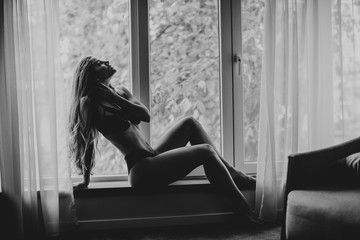 Beautiful young woman in lingerie sitting and looking outside the window in beautiful apartment - obrazy, fototapety, plakaty