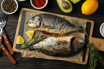 Cutting board with fried Dorado fishes and spice ingredients on wooden background, top view