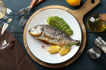 Plate with baked Dorado fish on garnished wooden background, top view