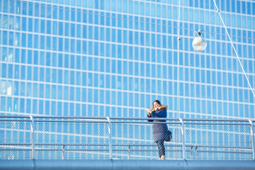 Fototapeta na wymiar Business woman at pedestrian bridge using her smartphone. A large city building serves as background.