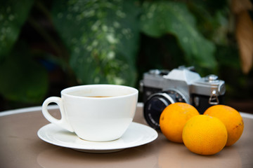 Obraz na płótnie Canvas a clear white ceramic cup and plate of black coffee with three orange and vintage camera on glass table with big green leaf background