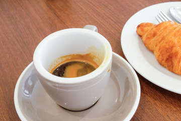Selective focus of white ceramic cup of espresso coffee shot place on nice and clean wooden table with fresh baked croissant blurred as background. Morning breakfast for refreshment or diet food.