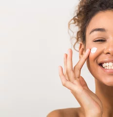 Poster Face care. Happy black woman applying moisturizer cream on cheek © Prostock-studio