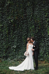 bride in a long white dress with a wedding bouquet along with a groom in a stylish suit after a wedding ceremony in front wall with green ivy