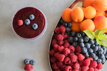 Fruit and berry smoothies and fresh apricots, raspberries and blueberries on an oval metal plate. Gray background.
