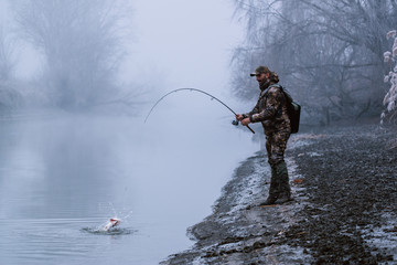 Fisher man fishing with spinning rod on a river bank at misty foggy winter, spin fishing, prey...