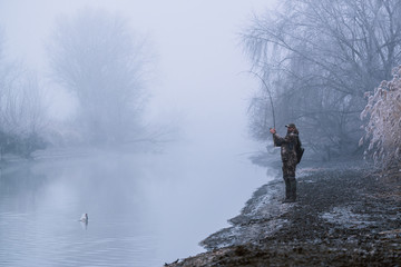 Fisher man fishing with spinning rod on a river bank at misty foggy winter, spin fishing, prey...