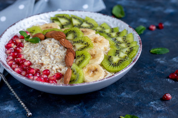 Breakfast consisting of oatmeal, nuts and fruits. Kiwi orange pomegranate almonds bananas decorate a plate. Healthy eating, on a dark background. Copy space.