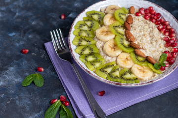 Breakfast consisting of oatmeal, nuts and fruits. Kiwi orange pomegranate almonds bananas decorate a plate. Healthy eating, on a dark background. Copy space.