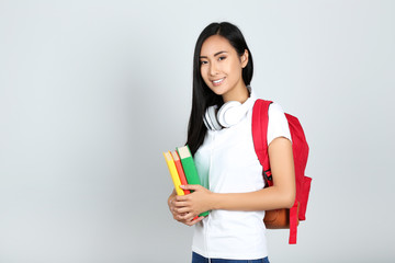 Young woman with books, headphones and backpack on grey background