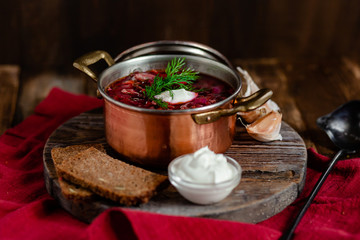 Freshly cooked homemade russian famous soup borsch. Served in cooper pan with garlic, sour cream and dill. Nourishing tasty meal. Rustic style, close up, wooden background, red tissue as decor. 
