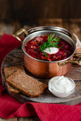 Freshly cooked homemade russian famous soup borsch. Served in cooper pan with garlic, sour cream and dill. Nourishing tasty meal. Rustic style, close up, wooden background, red tissue as decor. 