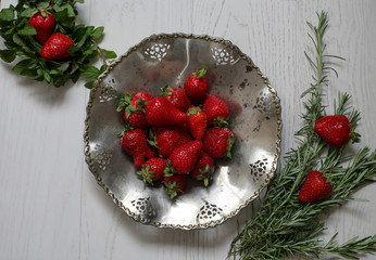 Top view of strawberries in a silver plate decorated with greenery on white wooden background.