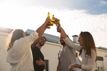 Joyful young friends drinking beer on balcony. Cheerful young people clinking beer bottles during...