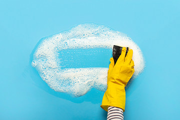 Hand in a yellow rubber glove holds a cleaning sponge and wipes a soapy foam on a blue background....