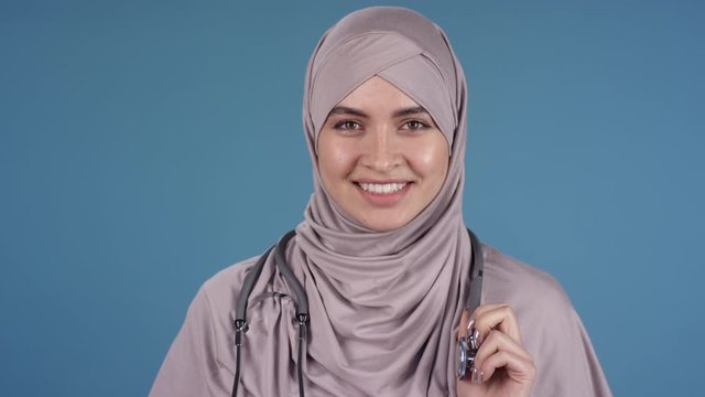 Dolly-out portrait shot of cheerful young female doctor wearing hijab and stethoscope around her neck standing isolated on blue background and smiling widely while looking at camera