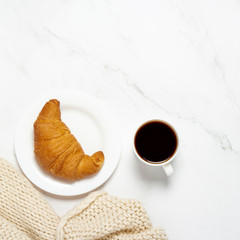Cup with coffee, croissant on a white plate and a knitted scarf on a marble table. Concept french breakfast, snack, work. Flat lay, top view