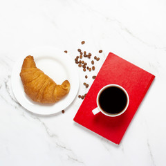 Cup with coffee on a book, croissant and coffee beans on a marble table. The concept of breakfast, freelance, work, french breakfast. Flat lay, top view