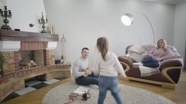 Portrait of playful Caucasian brunette girl dancing as her father playing ukulele and mother sitting on couch at the background. Cheerful cute child leaving shot to the left. Family, unity, leisure.