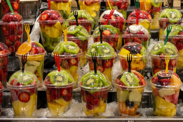 Fruit slices in transparent plastic cups with forks on a market counter: kiwi, orange, strawberry, banana. Refreshing bright fruits on a hot summer sunny day.