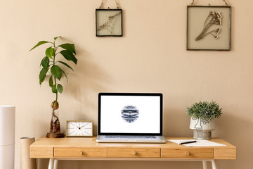 Stylish and creative wooden desk with laptop mock up screen, avocado plant office accessories, plant and gold clock. Beige background wall. Design home office interior.  Template.