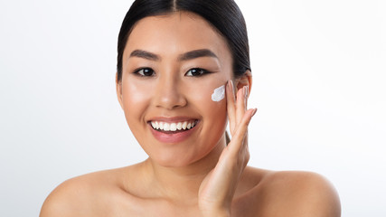 Morning routine. Asian girl applying cream on cheeks