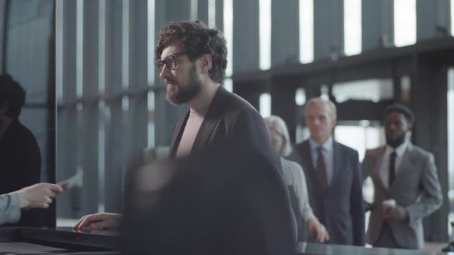 Waist-up arc shot of middle-aged Caucasian man in glasses, with full beard standing in queue at check-in counter in airport, giving passport and ticket to anonymous female agent, talking and nodding