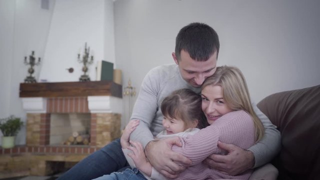 Young friendly Caucasian man, woman and little girl sitting on armchair and hugging. Smiling happy people posing indoors. Family, lifestyle, unity.