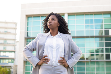 Confident young businesswoman with hands on waist. Low angle view of young African American businesswoman standing with hands on waist and looking away on street. Professional occupation concept