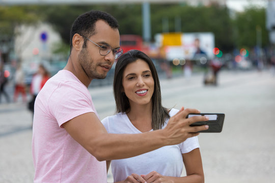 Cheerful couple posing for selfie on street. Happy multiracial friends taking self portrait with smartphone. Concept of self portrait