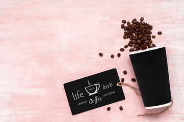 Black paper cup with coffee beans and a note card with text message on vintage pink wooden background. Top view. Flat lay