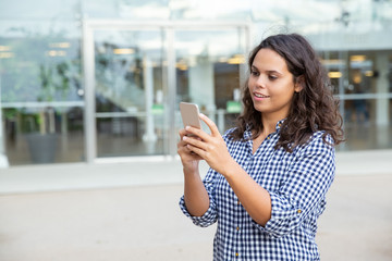 Smiling woman using smartphone on street. Beautiful cheerful young woman standing outside modern building and using mobile phone. Technology concept