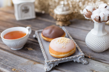 Donuts made of white milk chocolate, coated with glaze and nuts. Aristocratic tea party with porcelain cup on wooden table. Decorated with vases,cotton inflorescences,white sugar bowl.Gold background