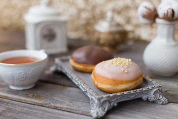 Donuts made of white milk chocolate, coated with glaze and nuts. Aristocratic tea party with porcelain cup on wooden table. Decorated with vases,cotton inflorescences,white sugar bowl.Gold background