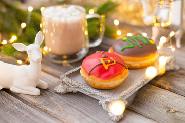 Obraz na płótnie Canvas Christmas chocolate donuts with Santa Claus icing and new year tree on ceramic stand. Glass cup of cocoa with marshmallows on wooden table. Spruce branch, present, cone, sugar bowl,white deer,garland