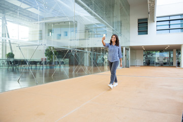 Fototapeta na wymiar Smiling woman walking and taking selfie with smartphone. Full length view of cheerful beautiful young woman walking inside modern building and using mobile phone. Technology concept
