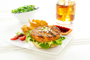 Open face fish patty sandwich on a square white plate with sides of tomatos, potato chips with a glass of ice tea and salad in the background