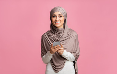Smiling islamic girl in headscarf holding smartphone and looking at camera