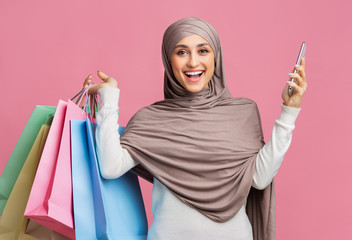 Joyful arabic woman in hijab holding smarphone and bright shopping bags
