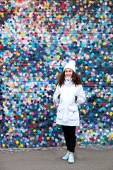 beautiful charming smiling young teen girl with curly loose hair looks posing against colorful wall in white jacket and hat