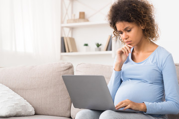 Confused afro expectant woman using laptop, buying goods for newborn