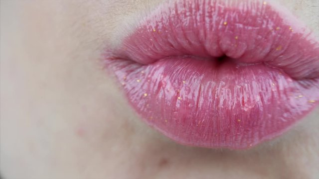 Woman is applying pink gloss on her lips and blowing a kiss. Close view of female lips with lipstick