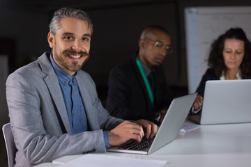 Obraz premium Smiling handsome manager sitting at table and looking at camera. Cheerful young man working with laptop in office at night. Business, working late concept
