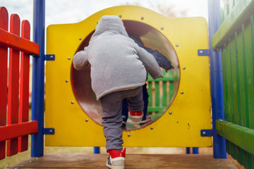 Small caucasian boy back view in the park on the playground in the yellow tunnel tube wearing winter coat in autumn day having fun playing walking